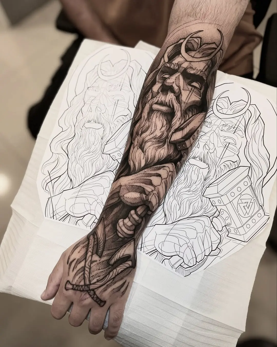 Tatuagem blackwork de Odin com elmo, barba e espada no antebraço